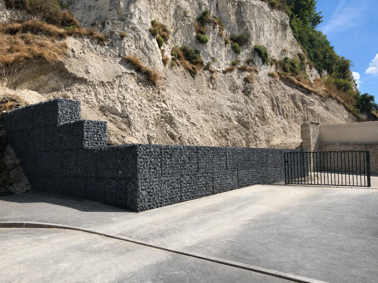 Mur de soutènement en gabions à mailles sombres, remplis de pierre concassée, au pied d'une falaise de calcaire.